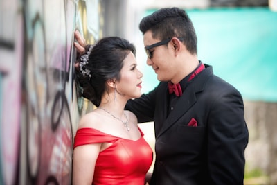 A couple stands close together in an intimate pose. The woman is wearing a red off-shoulder dress with elegant jewelry, her hair styled neatly. The man is dressed in a black suit with a red bow tie and pocket square, wearing sunglasses. They are positioned next to a graffiti-covered wall, with the man leaning against it, creating a romantic and stylish scene.