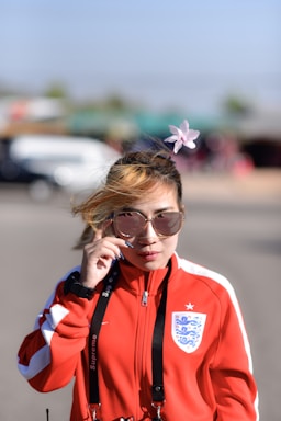 A person wearing a red and white sports jacket with an emblem, holding a pair of sunglasses. There is a flower hair accessory on the head and a lanyard around the neck. The background is blurred, indicating an outdoor setting with bright lighting.