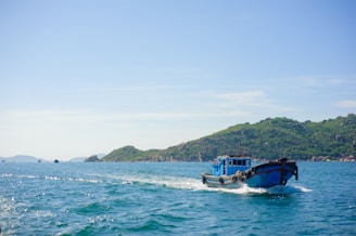 A bright blue fishing boat moves through clear turquoise waters, leaving behind a gentle wake. Lush green hills and rocky outcrops form a distant backdrop, bathed in soft sunlight under a clear blue sky.