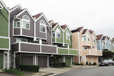 A row of colorful, modern townhouses with pointed roofs and large windows. Each house features unique pastel colors, including grey, green, and beige, accented by white trims. A car is parked on the street lining the houses, and small shrubs are visible in front of the buildings.