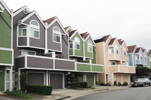 A row of colorful, modern townhouses with pointed roofs and large windows. Each house features unique pastel colors, including grey, green, and beige, accented by white trims. A car is parked on the street lining the houses, and small shrubs are visible in front of the buildings.