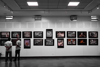 Two people stand in a gallery observing a collection of framed photographs displayed on a white wall. The ceiling is equipped with bright lights that illuminate the artwork. The room has a modern, clean design.