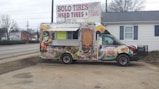A colorful food truck is parked in an outdoor area. It features vibrant graphics of food items like tacos and burritos, with a cartoon character on the side. The truck is advertising catering services with a phone number displayed. Behind the truck, there's signage for a tire shop, and the background includes a building and leafless trees.