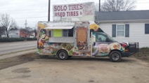 A colorful food truck is parked in an outdoor area. It features vibrant graphics of food items like tacos and burritos, with a cartoon character on the side. The truck is advertising catering services with a phone number displayed. Behind the truck, there's signage for a tire shop, and the background includes a building and leafless trees.