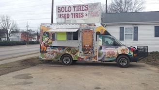 A colorful food truck is parked in an outdoor area. It features vibrant graphics of food items like tacos and burritos, with a cartoon character on the side. The truck is advertising catering services with a phone number displayed. Behind the truck, there's signage for a tire shop, and the background includes a building and leafless trees.