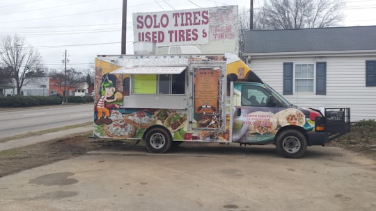 A colorful food truck is parked in an outdoor area. It features vibrant graphics of food items like tacos and burritos, with a cartoon character on the side. The truck is advertising catering services with a phone number displayed. Behind the truck, there's signage for a tire shop, and the background includes a building and leafless trees.
