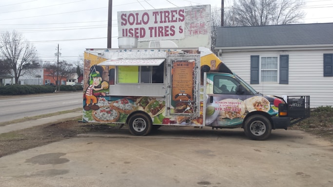 A colorful food truck is parked in an outdoor area. It features vibrant graphics of food items like tacos and burritos, with a cartoon character on the side. The truck is advertising catering services with a phone number displayed. Behind the truck, there's signage for a tire shop, and the background includes a building and leafless trees.
