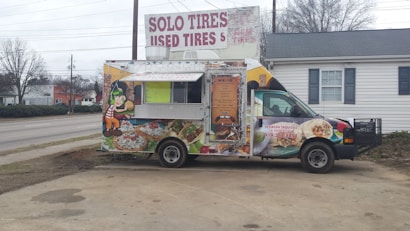 A colorful food truck is parked in an outdoor area. It features vibrant graphics of food items like tacos and burritos, with a cartoon character on the side. The truck is advertising catering services with a phone number displayed. Behind the truck, there's signage for a tire shop, and the background includes a building and leafless trees.