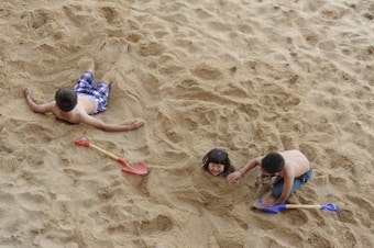 Three children are playing in the sand. Two boys, one wearing plaid shorts and the other in jeans, are using colorful shovels to dig and cover the girl in the sand, leaving only her head visible. The scene conveys a playful mood in a beach or sandpit area.