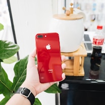 A hand holds a red iPhone with the Apple logo, positioned in a bright room next to a decorative pot on a wooden stand. In the background, a bottle of soda and green leaves can be seen.