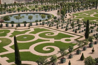 An intricately designed formal garden featuring geometric patterns of trimmed grass bordered by stone pathways. A circular body of water, around which are symmetrically planted trees and shrubs, adds to the organized beauty. The garden displays a harmonious blend of nature and design.