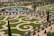 An intricately designed formal garden featuring geometric patterns of trimmed grass bordered by stone pathways. A circular body of water, around which are symmetrically planted trees and shrubs, adds to the organized beauty. The garden displays a harmonious blend of nature and design.
