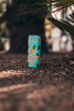 A colorful beverage can is placed on the ground, surrounded by an outdoor setting with palm leaves partially visible in the background. The can is designed with tropical motifs, such as pineapples and leaves, and features a vibrant turquoise color.