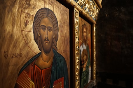 Iconography featuring a religious depiction with intricate details. The foreground shows a figure with a serene expression, surrounded by a golden halo and detailed with Greek lettering. The background includes another religious image in a richly decorated frame.