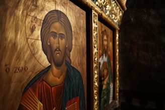 Iconography featuring a religious depiction with intricate details. The foreground shows a figure with a serene expression, surrounded by a golden halo and detailed with Greek lettering. The background includes another religious image in a richly decorated frame.