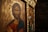 Iconography featuring a religious depiction with intricate details. The foreground shows a figure with a serene expression, surrounded by a golden halo and detailed with Greek lettering. The background includes another religious image in a richly decorated frame.