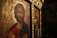 Iconography featuring a religious depiction with intricate details. The foreground shows a figure with a serene expression, surrounded by a golden halo and detailed with Greek lettering. The background includes another religious image in a richly decorated frame.