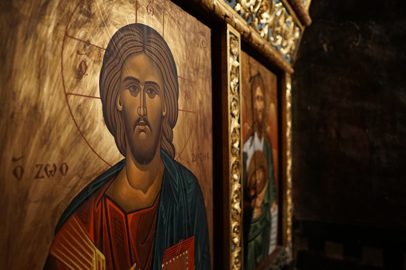 Iconography featuring a religious depiction with intricate details. The foreground shows a figure with a serene expression, surrounded by a golden halo and detailed with Greek lettering. The background includes another religious image in a richly decorated frame.