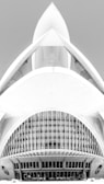 A black and white architectural photograph featuring a modern building design with a futuristic appearance. The structure has a symmetrical design with curved lines, large arches, and intricate detailing, resembling a spaceship or an abstract sculpture. The central feature includes narrow vertical windows and a smooth concrete facade.