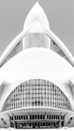 A black and white architectural photograph featuring a modern building design with a futuristic appearance. The structure has a symmetrical design with curved lines, large arches, and intricate detailing, resembling a spaceship or an abstract sculpture. The central feature includes narrow vertical windows and a smooth concrete facade.