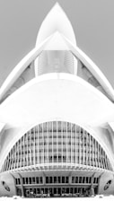 A black and white architectural photograph featuring a modern building design with a futuristic appearance. The structure has a symmetrical design with curved lines, large arches, and intricate detailing, resembling a spaceship or an abstract sculpture. The central feature includes narrow vertical windows and a smooth concrete facade.