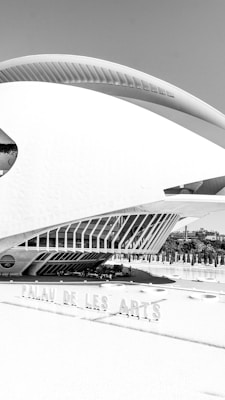 A modern architectural structure with sleek, futuristic lines and bold geometric patterns. The building features large windows and a sweeping curved roof, with a surrounding area that includes landscaping and a reflecting pool. The structure is prominently labeled 'Palau de les Arts.'