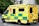 A large, yellow and green emergency ambulance is parked on a city street. The vehicle is part of the London Ambulance Service, as indicated by the text and logos on its side. It features red and yellow chevron patterns on the back doors and displays several identification numbers and emergency contact information.