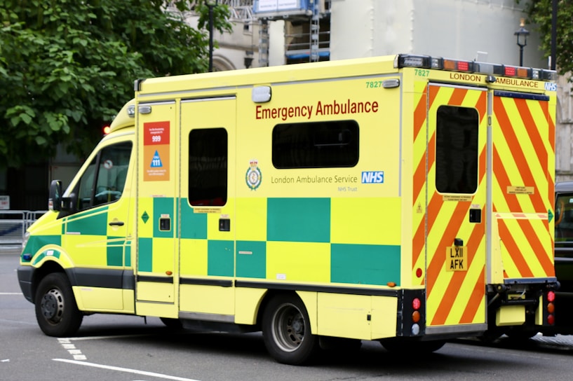 A large, yellow and green emergency ambulance is parked on a city street. The vehicle is part of the London Ambulance Service, as indicated by the text and logos on its side. It features red and yellow chevron patterns on the back doors and displays several identification numbers and emergency contact information.