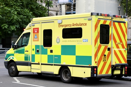 A large, yellow and green emergency ambulance is parked on a city street. The vehicle is part of the London Ambulance Service, as indicated by the text and logos on its side. It features red and yellow chevron patterns on the back doors and displays several identification numbers and emergency contact information.