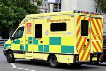 A large, yellow and green emergency ambulance is parked on a city street. The vehicle is part of the London Ambulance Service, as indicated by the text and logos on its side. It features red and yellow chevron patterns on the back doors and displays several identification numbers and emergency contact information.