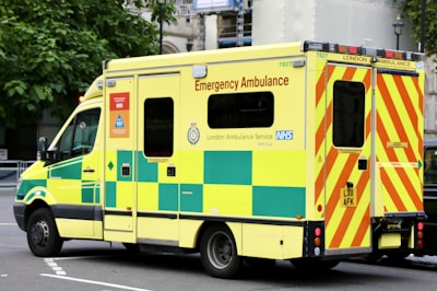A large, yellow and green emergency ambulance is parked on a city street. The vehicle is part of the London Ambulance Service, as indicated by the text and logos on its side. It features red and yellow chevron patterns on the back doors and displays several identification numbers and emergency contact information.