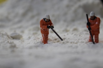Two miniature figures dressed in orange work gear and helmets appear to be shoveling or working in a field of a white substance, resembling snow or sand.