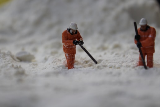 Two miniature figures dressed in orange work gear and helmets appear to be shoveling or working in a field of a white substance, resembling snow or sand.