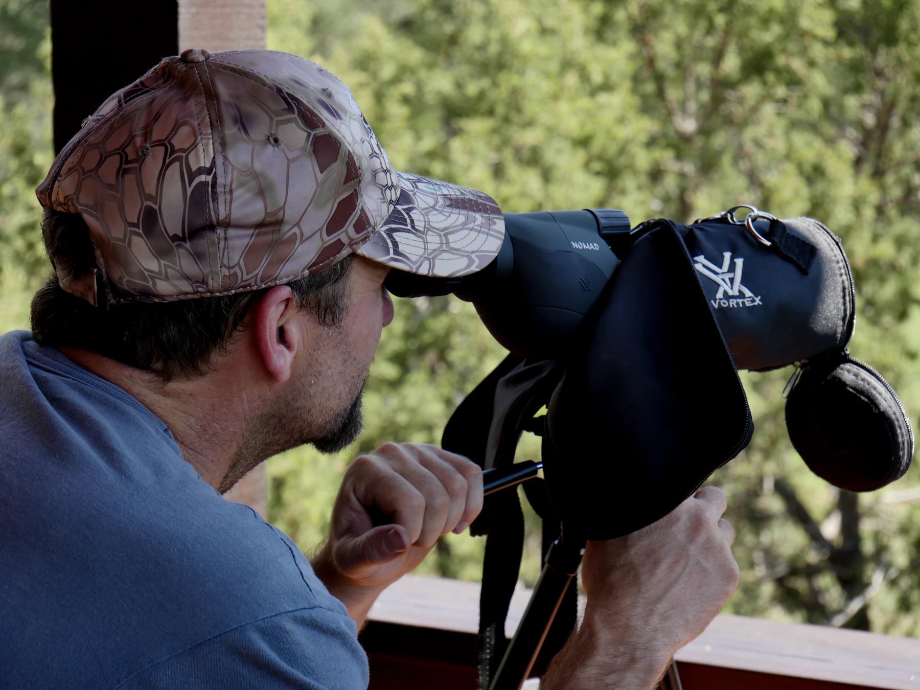A man in a cap looking through an angled spotting scope from a wooden deck, trees behind