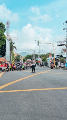 A busy urban intersection with multiple motorcyclists lined up on a road. A person, possibly a traffic officer, stands in the middle directing the flow. In the background, there are buildings, trees, and telephone wires against a partly cloudy sky.