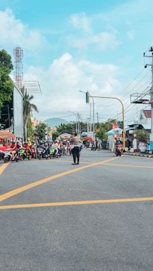 A busy urban intersection with multiple motorcyclists lined up on a road. A person, possibly a traffic officer, stands in the middle directing the flow. In the background, there are buildings, trees, and telephone wires against a partly cloudy sky.