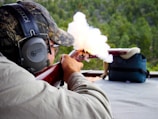 A person is aiming and firing a rifle at a shooting range, with smoke visibly coming out of the gun. The individual is wearing protective earmuffs and a camouflage hat.