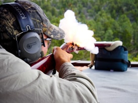 A person is aiming and firing a rifle at a shooting range, with smoke visibly coming out of the gun. The individual is wearing protective earmuffs and a camouflage hat.