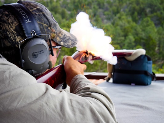 A person is aiming and firing a rifle at a shooting range, with smoke visibly coming out of the gun. The individual is wearing protective earmuffs and a camouflage hat.