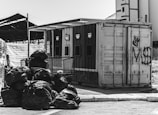A black and white image depicting a recycling facility with separate sections labeled for different materials. A pile of garbage bags is situated in the foreground, while various graffiti is present on the container in the background.