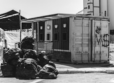 A black and white image depicting a recycling facility with separate sections labeled for different materials. A pile of garbage bags is situated in the foreground, while various graffiti is present on the container in the background.