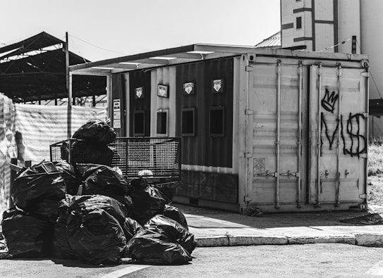 A black and white image depicting a recycling facility with separate sections labeled for different materials. A pile of garbage bags is situated in the foreground, while various graffiti is present on the container in the background.