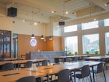 A modern cafeteria interior featuring wooden tables and black chairs arranged neatly in rows. The ceiling is adorned with industrial-style lighting with exposed pipes. Large windows allow natural light to flood the space, offering views of nearby buildings. A counter with a geometric herringbone pattern and a neon sign is present at the back, along with potted plants.