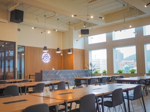 A modern cafeteria interior featuring wooden tables and black chairs arranged neatly in rows. The ceiling is adorned with industrial-style lighting with exposed pipes. Large windows allow natural light to flood the space, offering views of nearby buildings. A counter with a geometric herringbone pattern and a neon sign is present at the back, along with potted plants.