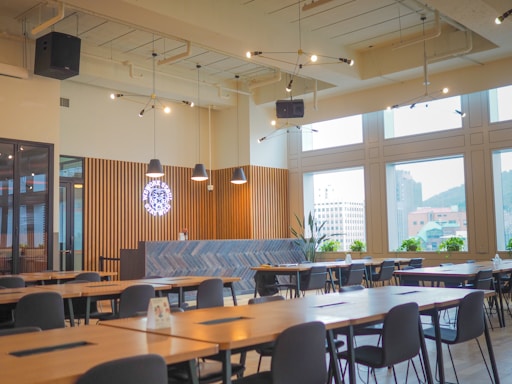 A modern cafeteria interior featuring wooden tables and black chairs arranged neatly in rows. The ceiling is adorned with industrial-style lighting with exposed pipes. Large windows allow natural light to flood the space, offering views of nearby buildings. A counter with a geometric herringbone pattern and a neon sign is present at the back, along with potted plants.