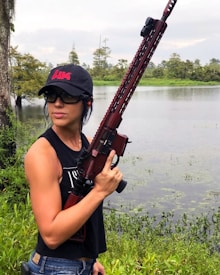 A person is standing near a body of water holding a large rifle. They are wearing a black sleeveless shirt, blue jeans, and a black cap with red lettering. The background includes a lake, trees, and a cloudy sky.