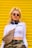 A person with long blonde hair tied up in a high ponytail wears sunglasses and large earrings. They are dressed in a white long-sleeve crop top with the word 'MIAMI' on it, camo pants, a crossbody bag with a brown and beige pattern, and headphones around their neck. A yellow shutter serves as the background.