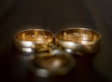 Two gold wedding rings are placed side by side. The rings have intricate engravings on the inside, with soft lighting creating a warm ambiance. The background is blurred, focusing attention on the rings.