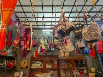 A variety of colorful bags and fabrics are hanging from a ceiling grid in a shop. The ceiling has ropes and hooks holding the items. Below are wooden cabinets with more textiles and accessories displayed. The shop is vibrant with an assortment of patterns and colors.