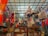A variety of colorful bags and fabrics are hanging from a ceiling grid in a shop. The ceiling has ropes and hooks holding the items. Below are wooden cabinets with more textiles and accessories displayed. The shop is vibrant with an assortment of patterns and colors.